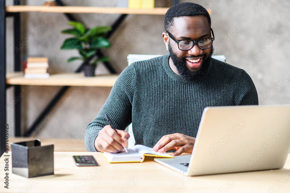 Cheerful African-American male student or worker in glasses is watching online lectures or webinars and writing notes in a notebook